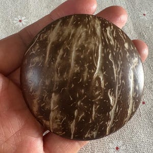 Op de afbeelding: Een rond, donkerbruin object met een natuurlijk, getextureerd patroon van lichtbruine strepen. Het object wordt in een hand gehouden, wat de grootte en vorm laat zien. Het oppervlak heeft een gepolijste uitstraling, wat de houtnerf benadrukt.