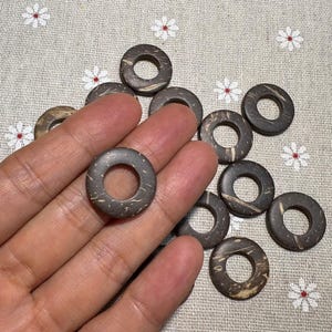 Puede incluir: Una colecci&oacute;n de cuentas circulares de color marr&oacute;n oscuro con un agujero en el centro. Las cuentas tienen un aspecto natural y texturizado, y se sostienen en una mano. El fondo es una tela clara con un estampado floral.