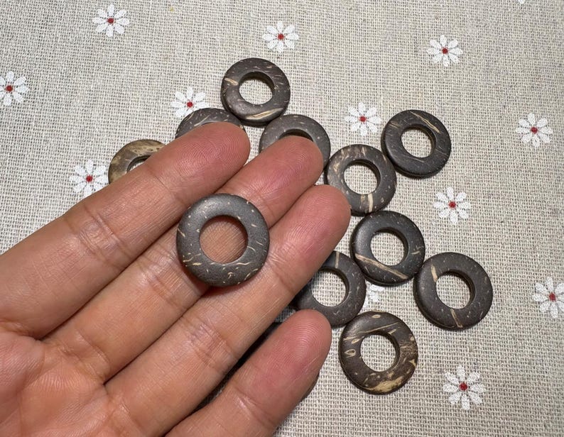 Puede incluir: Una colecci&oacute;n de cuentas circulares de color marr&oacute;n oscuro con un agujero en el centro. Las cuentas tienen un aspecto natural y texturizado, y se sostienen en una mano. El fondo es una tela clara con un estampado floral.