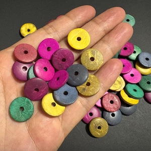 May include: A hand displays a selection of colourful, round wooden beads. The beads are in shades of pink, yellow, green, and blue. Each bead has a central hole, and they appear to be made from natural wood.