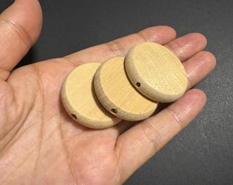 May include: Three round, unfinished wooden beads with a light tan color. Each bead has a small hole drilled through the center. The beads are held in a hand against a dark background.