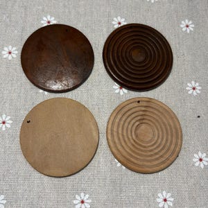 May include: Four wooden coasters in the image. Two are dark brown, one is plain, and the other has concentric circles carved into it. The other two are light brown, one plain and the other with a carved concentric circle design. Each coaster has a small hole.