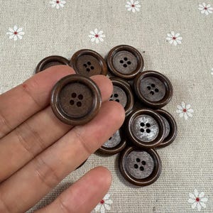 Puede incluir: Un conjunto de botones de madera marrón oscuro, cada uno con cuatro agujeros. Los botones son redondos con un borde ligeramente elevado. Los botones se muestran sobre una tela beige claro con un estampado floral.