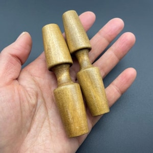 Puede incluir: Dos tapones de botella de madera de color marrón claro con una parte superior cónica y un cuerpo cilíndrico. Los tapones se sostienen en una mano sobre un fondo oscuro. La veta de la madera es visible en la superficie de los tapones.