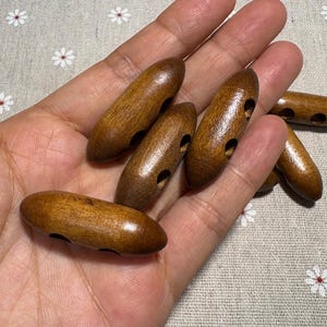 Puede incluir: Un conjunto de botones de madera ovalados en varios tonos de marrón. Cada botón tiene dos agujeros para coser. Los botones son lisos y tienen un aspecto de veta de madera natural. Se muestran sobre un fondo claro.