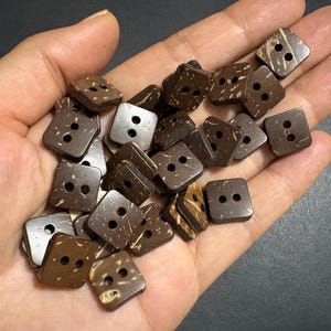 May include: A collection of small, square, dark brown buttons with rounded corners. Each button features a two-hole design in the center and a natural, mottled texture across the surface, showing variations in color and pattern on each piece.