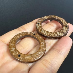 May include: Two brown, circular wooden beads with a speckled pattern and three small holes. The beads are shown against a dark background. These beads are suitable for jewelry making and craft projects.
