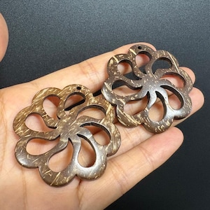 May include: Two brown, flower-shaped pendants made from coconut shell. Each pendant has a series of curved cutouts, creating a delicate, open design. The pendants are held in a hand against a dark background.