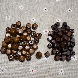 May include: Two piles of wooden beads in shades of brown. The cube-shaped beads have rounded edges and a central hole. They are scattered on a light grey fabric with a white floral pattern.