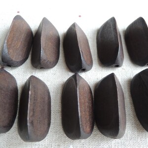 Può includere: Sono mostrate dieci perline di legno a forma di goccia, di colore marrone scuro. Ogni perlina ha un foro per l'infilatura. Le perline hanno una finitura liscia e lucida. Sono disposte su una superficie chiara.