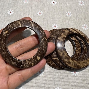 Puede incluir: Un conjunto de cuatro pulseras de c&aacute;scara de coco marr&oacute;n oscuro. Las pulseras tienen forma circular con un agujero en el centro. El patr&oacute;n natural de la veta de la madera es visible en la superficie de las pulseras.