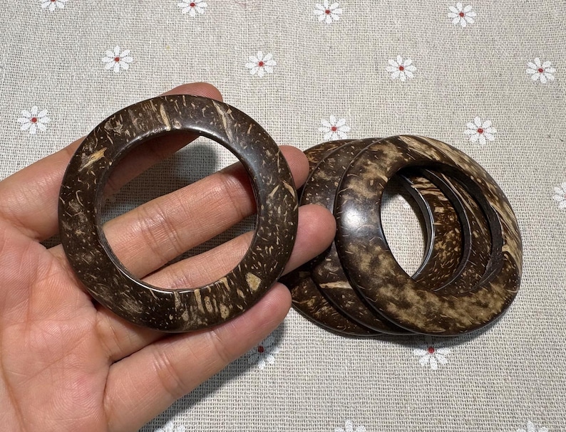 Puede incluir: Un conjunto de cuatro pulseras de c&aacute;scara de coco marr&oacute;n oscuro. Las pulseras tienen forma circular con un agujero en el centro. El patr&oacute;n natural de la veta de la madera es visible en la superficie de las pulseras.