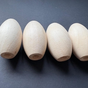 May include: Four unfinished wooden oval-shaped beads. Each bead is a light beige color with visible wood grain. The beads have a cylindrical hole drilled through the center, and are arranged in a row.