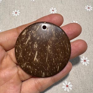 Könnte beinhalten: Ein runder, dunkelbrauner Anhänger aus Kokosnussschale, gehalten in einer Hand. Der Anhänger hat eine natürliche, strukturierte Oberfläche mit helleren Fasern und ein kleines Loch oben zum Auffädeln. Das Gesamtthema ist natürlich und erdig.