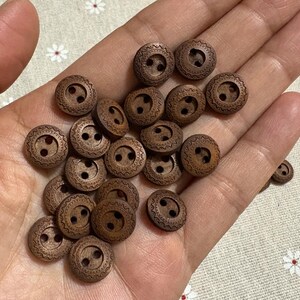 May include: A selection of small, round, wooden buttons, each with two holes and a textured border. The buttons are a warm brown hue and appear handmade. They are held in a person's hand.
