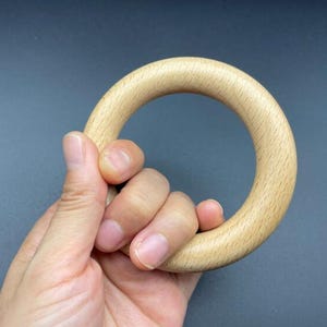 May include: A light-colored wooden ring, held in a hand. The ring is smooth and circular, with a natural wood grain visible. The background is a solid, dark gray color.