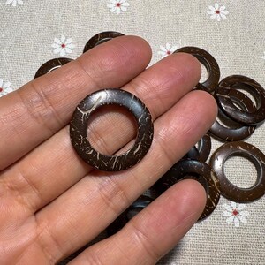 Puede incluir: Un conjunto de anillos de coco marr&oacute;n oscuro con un aspecto natural y texturizado. Los anillos tienen forma circular con un agujero en el centro. Los anillos se sostienen en una mano y son adecuados para manualidades y bisuter&iacute;a.