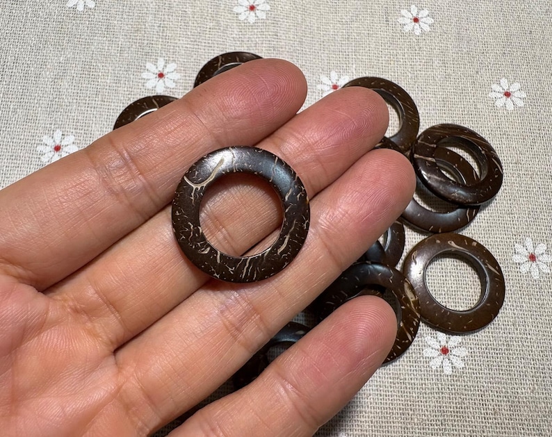 Puede incluir: Un conjunto de anillos de coco marr&oacute;n oscuro con un aspecto natural y texturizado. Los anillos tienen forma circular con un agujero en el centro. Los anillos se sostienen en una mano y son adecuados para manualidades y bisuter&iacute;a.