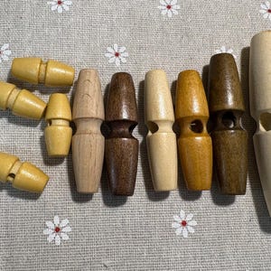 Może przedstawiać: Kolekcja drewnianych guzików typu toggle w różnych odcieniach brązu i beżu. Guziki są cylindryczne z otworem do szycia. Niektóre mają kształt klepsydry. Ułożone na szarej tkaninie z białymi wzorami kwiatowymi.