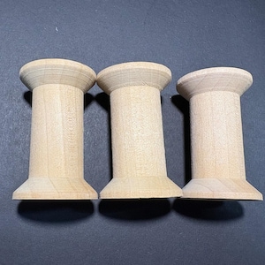 Può includere: Tre rocchetti di legno non rifiniti con venatura naturale. Ogni rocchetto ha una base e una parte superiore larghe, collegate da un corpo cilindrico. I rocchetti sono disposti in fila, mostrando il loro design semplice e classico.
