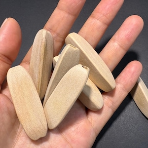 Puede incluir: Un conjunto de espigas de madera sin terminar, de color claro, cada una con forma rectangular redondeada. Las espigas se sostienen en una mano abierta sobre un fondo oscuro, mostrando la veta natural de la madera y su textura suave.