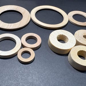 Op de afbeelding: Een verzameling onafgewerkte houten ringen in verschillende maten en vormen. De ringen zijn lichtbruin van kleur en lijken van natuurlijk hout te zijn. Sommige ringen zijn plat, andere cilindrisch. De objecten zijn op een donkere ondergrond geplaatst.