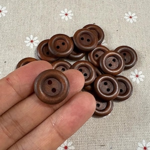 Puede incluir: Un conjunto de botones de madera marrón oscuro, cada uno con dos agujeros. Los botones son redondos con un borde ligeramente elevado, colocados sobre una tela beige claro con un estampado floral. Los botones tienen un estilo natural y rústico.