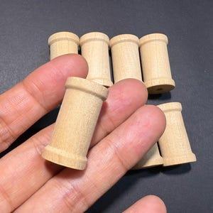 Può includere: Una collezione di piccole bobine di legno non rifinite. Le bobine sono cilindriche con una base e una parte superiore leggermente svasate. Sono realizzate in legno di colore chiaro e sembrano nuove. Le bobine sono disposte su una superficie scura.