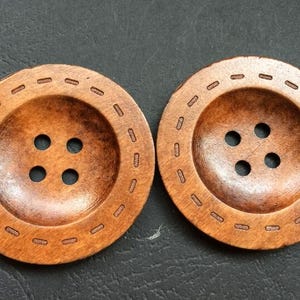Puede incluir: Dos botones grandes y redondos de madera con un acabado marrón cálido. Cada botón tiene cuatro agujeros y un borde decorativo cosido. Los botones están sobre un fondo oscuro.