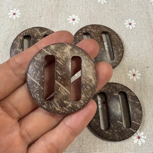 Puede incluir: Cuatro botones redondos de coco marrón oscuro con dos ranuras rectangulares. Los botones tienen un aspecto natural y texturizado y se sostienen en una mano. El fondo es una tela beige claro con pequeños estampados de flores blancas.