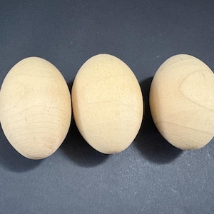 Może przedstawiać: Trzy niedokończone drewniane kształty jaj, ułożone poziomo na ciemnym tle. Jajka mają jasny, naturalny kolor drewna z widocznymi wzorami słojów. Są gładkie i owalne, idealne do projektów rzemieślniczych.