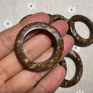 Puede incluir: Una mano sostiene varios anillos circulares marrones hechos de un material natural, posiblemente madera o c&aacute;scara de coco. Los anillos tienen una superficie texturizada con una combinaci&oacute;n de colores marr&oacute;n oscuro y beige. El fondo es una tela clara con un estampado floral.