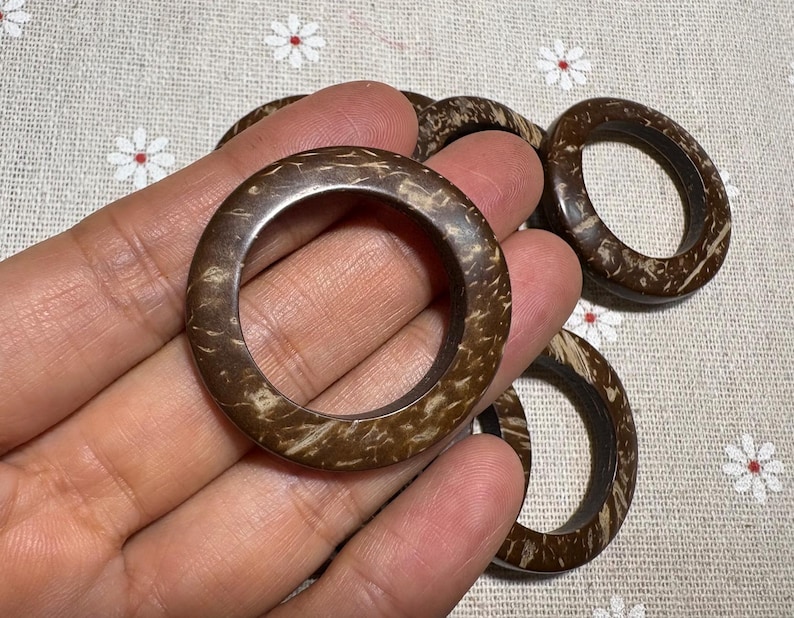 Puede incluir: Una mano sostiene varios anillos circulares marrones hechos de un material natural, posiblemente madera o c&aacute;scara de coco. Los anillos tienen una superficie texturizada con una combinaci&oacute;n de colores marr&oacute;n oscuro y beige. El fondo es una tela clara con un estampado floral.