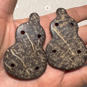 Puede incluir: Dos colgantes en forma de calabaza con tres agujeros cada uno. Los colgantes son de color marrón oscuro con vetas marrones más claras y tienen una superficie texturizada. Los colgantes están hechos de un material natural.