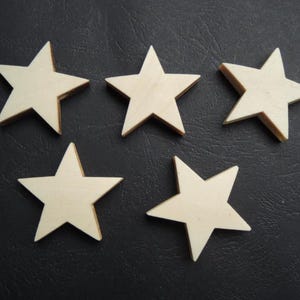 May include: Five wooden star cutouts with a natural wood grain finish. The stars are arranged on a dark, textured surface. Each star has a five-point design, ideal for crafts and decorations.