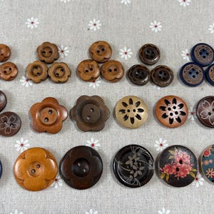 May include: A collection of various wooden buttons in shades of brown, tan, and blue. The buttons feature different shapes, including round, flower, and carved designs. Some have floral patterns and decorative details.
