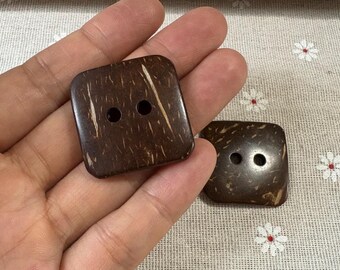 Könnte beinhalten: Zwei quadratische, dunkelbraune Kokosnussschalenknöpfe mit je zwei Löchern. Die Knöpfe haben eine natürliche, strukturierte Oberfläche mit helleren braunen Streifen. Ein Knopf wird in einer Hand gehalten.