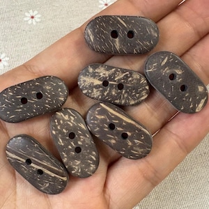 Può includere: Una collezione di bottoni ovali in guscio di cocco, marrone scuro, con due fori ciascuno. I bottoni hanno un aspetto naturale e strutturato con venature marroni più chiare. Ideali per cucito e progetti artigianali.
