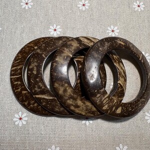 Puede incluir: Cuatro brazaletes de concha de coco marr&oacute;n oscuro con patrones de vetas de madera natural. Los brazaletes est&aacute;n apilados, mostrando su forma circular y textura org&aacute;nica. El fondo es una tela gris claro con un estampado floral.