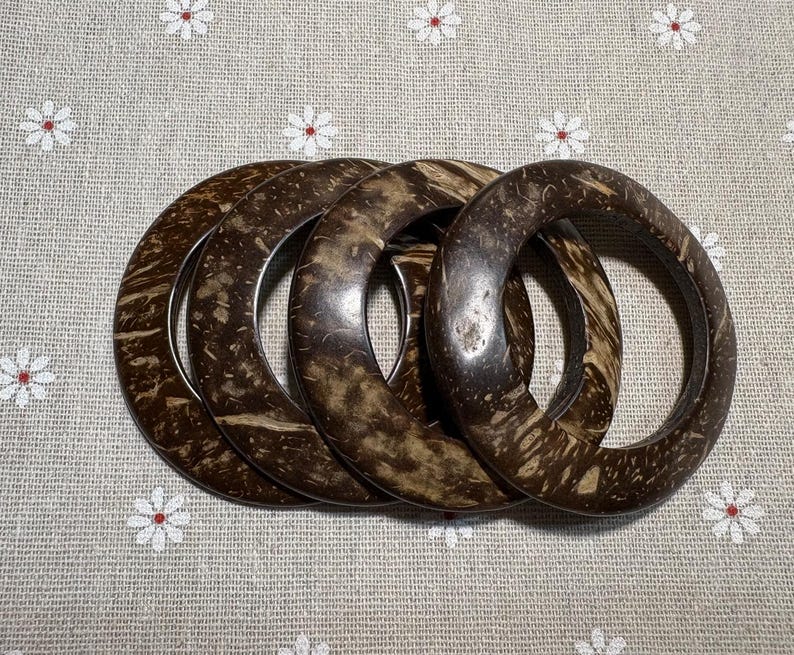 Puede incluir: Cuatro brazaletes de concha de coco marr&oacute;n oscuro con patrones de vetas de madera natural. Los brazaletes est&aacute;n apilados, mostrando su forma circular y textura org&aacute;nica. El fondo es una tela gris claro con un estampado floral.