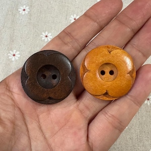 May include: Two wooden buttons with a flower design. One button is dark brown, the other is light orange. Each button has two holes for sewing. The buttons are round and have a carved floral pattern around the edges.