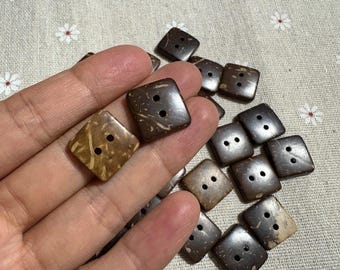 Könnte beinhalten: Eine Sammlung quadratischer Knöpfe mit zwei Löchern aus dunkelbraunem Material. Einige Knöpfe haben eine hellere, hellbraune Farbe. Die Knöpfe liegen auf einem hellen Stoff mit einem floralen Muster.