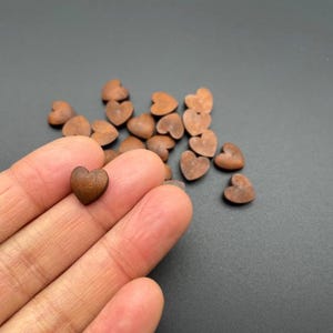 May include: A selection of small, heart-shaped wooden beads in shades of brown. One bead is held in a hand, with others scattered on a grey surface. The beads have a smooth, polished appearance, suitable for crafting or jewellery making.
