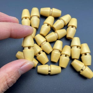 May include: A collection of light-colored wooden toggle clasps. Each clasp has a cylindrical shape with a rounded top and bottom, and a hole for threading. The clasps are a natural wood color and are suitable for various craft projects.