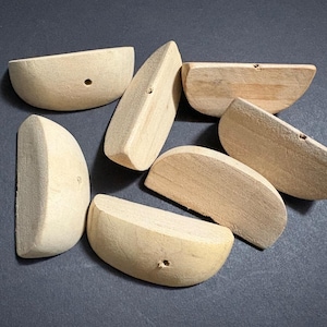 Puede incluir: Una colección de piezas de madera sin terminar para manualidades, cada una con una forma única y un pequeño agujero. Las piezas son de color claro y parecen estar hechas de madera natural, dispuestas sobre una superficie oscura.