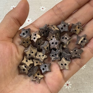 May include: A collection of star-shaped buttons in varying shades of brown and tan. Each button has two holes for sewing. The buttons are made of a natural material, possibly wood or coconut shell.