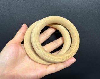 May include: Two light-colored wooden rings, possibly for a baby toy or craft project, are held in a person's hand against a dark background. The rings are smooth and appear to be made of unfinished wood.