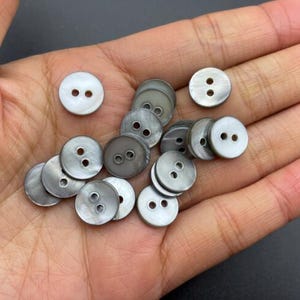 Puede incluir: Un conjunto de botones redondos, cada uno con dos agujeros, en tonos blancos y grises. Los botones, hechos de un material nacarado, se muestran en una mano abierta.