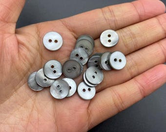 Könnte beinhalten: Eine Sammlung runder Knöpfe mit jeweils zwei Löchern in Weiß- und Grautönen. Die Knöpfe aus Perlmuttmaterial werden in einer offenen Hand gezeigt.