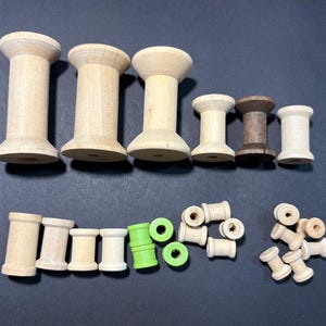 Puede incluir: Una colección de carretes de madera de varios tamaños y colores. Los carretes varían de madera clara a marrón oscuro, algunos pintados de verde. Están dispuestos sobre una superficie oscura, mostrando sus diferentes formas y tamaños.